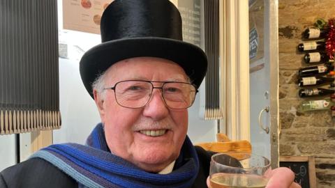The image shows a man indoors, wearing a top hat and glasses. He is smiling at the camera and holding a glass. Behind him is a window with grey blinds, and a door. 