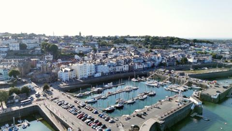 An aerial photo of the harbour and St Peter Port on a bright day. There are many yacht and tenders moored up and the harbour car park looks full.