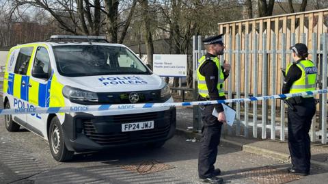 Two police officers stand behind a line of police tape beside a police van. In the background is an NHS sign saying "Hunter Street Health and Care Centre".