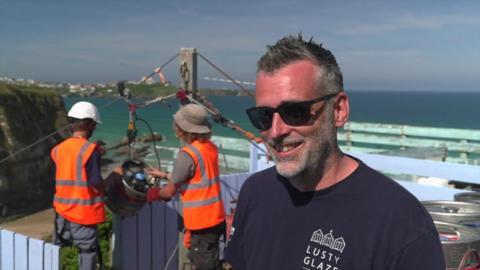 A photo of Robin Parris. It shows two people behind him in hi-vis jackets pushing the beer keg down the zipline. Mr Parris is in the forefront wearing sunglasses and a T-shirt saying Lusty Glaze