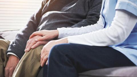 A nurse sits next to an elderly patient with her right hand on his left hand, which is resting on his left knee.