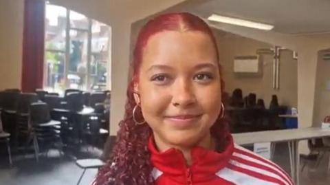 A girl with red hair and tracksuit top smiles for the camera 