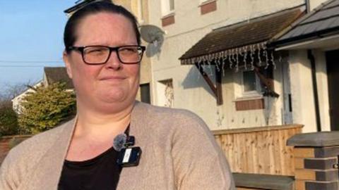 Maria Michaels has dark hair tied back and is wearing a black framed glasses, a black top and beige cardigan. She is stood outside a cream coloured rendered house and looking into the distance.