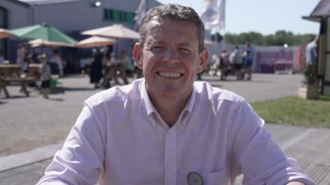First Minister Rhun ap Iorwerth is sat down looking into the camera. He has short light brown hair, and is wearing a light, lilac shirt and is smiling. In the background there are festival-goers of the Urdd Eisteddfod, some are walking while others are sat on benches under parasols. There is also a large grey building far in the background and there are white flags or signs flapping in the wind.