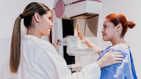 A female doctor doing mammogram exam on patient at hospital - stock photo. Both women have their hair tied back. The doctor is wearing a white lab coat and patient is wearing a blue gown.