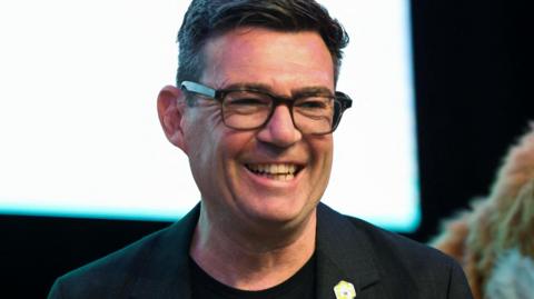 Newly elected Makerfield MP Andy Burnham smiles after winning the Makerfield by-election. He has short brown hair and is wearing a black blazer and t-shirt. He wears dark-framed glasses.