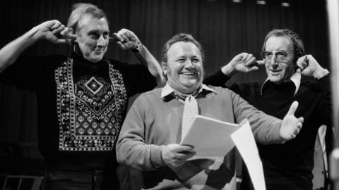 A black and white image of three smiling men, performing a show. The man on the left is tall, wearing a black jumper with an embroidered pattern. He has his fingers in his ears. The man in the middle holds a script is smiling, wearing a jumper, shirt and a tie. The man on the right also has his fingers in his ears, and wears large glasses, a dark jumper and a shirt with a large collar.
