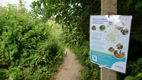 The image contains large green bushes on either side of a path which snakes down further up ahead. There is also a stone pillar with a sign attached to it talking about new outdoor activities South Farnham needs.