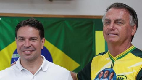 Alexandre Ramagem, wearing a white Polo shirt and standing in front of a Brazilian flag, smiles at the camera. Next to him, with his hand placed on Ramagem's shoulder, is Jair Bolsonaro. He is wearing a yellow-and-green football shirt