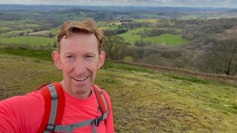 Karl Thompson smiling at the camera wearing a coral long sleeve running top and a red back-pack with hills and greenery in the background.