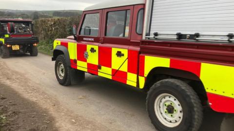 Emergency fire vehicles parked on a single track road. They have yellow and red markings on the side of the 4x4 vehicles. 