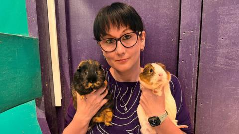 A woman with short black hair and glasses holds two guinea pigs while looking at the camera