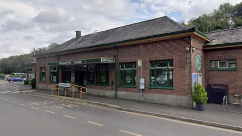 Outside of Okehampton railway station in Devon. It is a red brick building with green signs with Okehampton Station written on. It has a bus stop outside the front of the station. A blue car is parked in the distance.