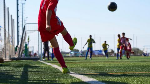 A young footballer in a red kit takes a corner and a number of players are waiting in the box to try and get to the ball.