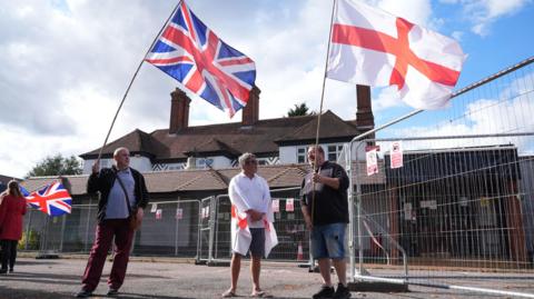Three men standing outside a large house that is surrounded by metal fencing. The three men are wearing polo shirts, two wearing shorts and two are holding large flags - one is the Union Jack the other is the St George's Cross.