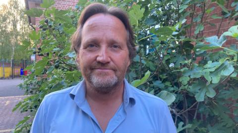 Councillor Matt Dormer is standing outside, wearing a blue shirt. Behind him is a bush, and the road.