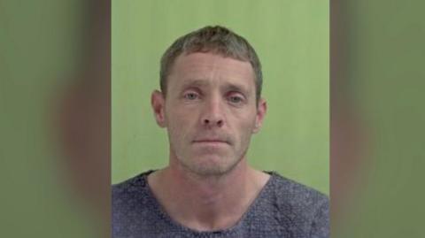 A middle-aged man wearing a grey jumper looks somberly at the camera, in front of a green background in a police mugshot.