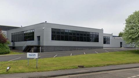 The grey building has lots dark glass windows. It has a flat roof and an empty car park in the foreground with grass and a tree.