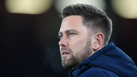 Des Buckingham looks slightly to his left with a bemused look. He wears a navy hooded coat.
