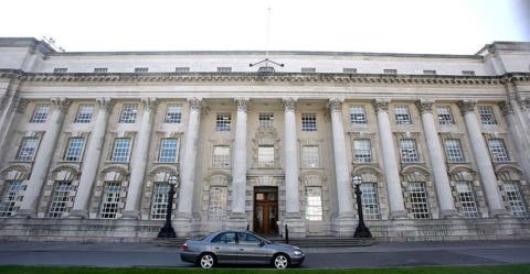 A front view of an old, stately-looking building. It is four storeys high and there are long grey columns going up three storeys. In the middle on the ground floor is a brown, wooden double door as an entrance. In front of the door is a single parked car, a grey four-door. 