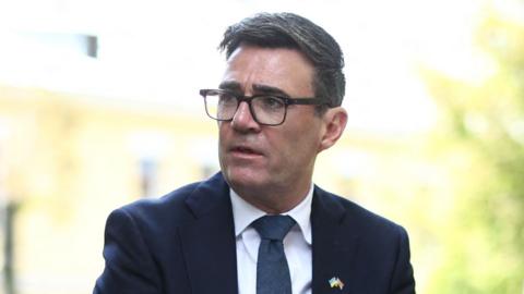 Andy Burnham wearing a black suit and white shirt and glasses, looking to the left of the frame of the photograph. The picture is a close up with most of Burnham out of shot bar his head and shoulders.