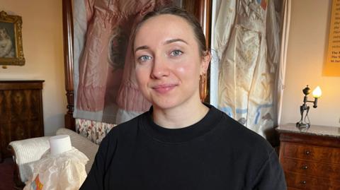 Naomi Deller a woman wearing a black long sleeved T-shirt with brown hair which is pulled back off her face. She is looking directly at the camera and smiling.