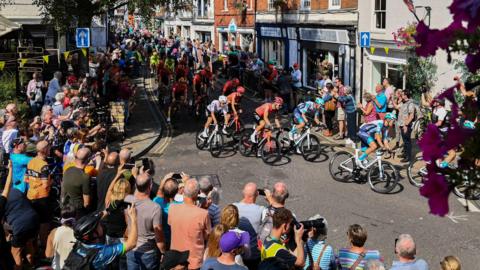 A large group of cyclists riding through a town with crowds cheering them on either side of the street. Some people are taking pictures of the cyclists.