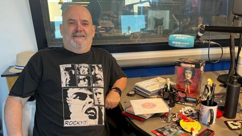 Glyn is seated in a radio studio, wearing a black 'Rocky!!' graphic T-shirt, surrounded by Rocky Horror memorabilia including figurines, books, and a mug on the table. Studio equipment and monitors are visible through a window behind them.