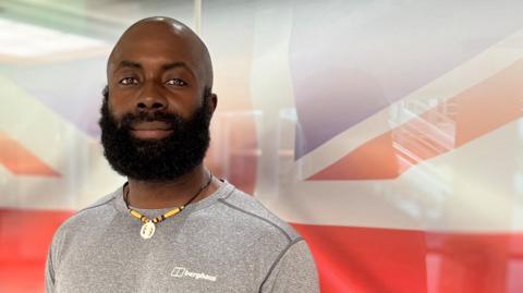 A man stands in front of a Union flag backdrop at the Hull 4 Heroes headquarters in Princes Quay, Hull. He wears a grey T-shirt and beaded necklace. He has a bald head a large black beard. He is smiling.