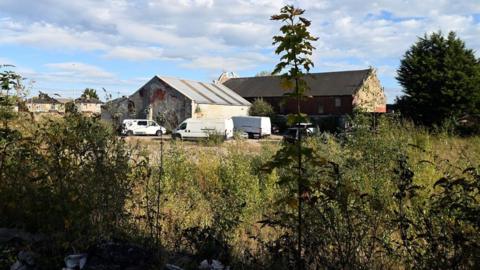 A group of derelict buildings are in the distance with a number of parked white vans next to them. In the foreground of the image is overgrown weeds and shrubs