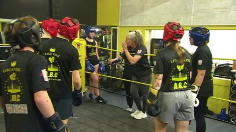 Heidi Ogden, leaning on the ropes of a boxing ring talking to some young people in the ring wearing different coloured gloves and head guards.