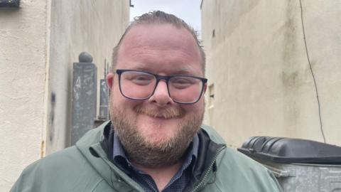 An image of William Banks, standing in the entrance to the alley way, there is a silver coloured metal gate behind his left shoulder and a silver coloured industrial bin with a black lid over his right shoulder. William is a balding man with a beard, he is smiling and wearing blue rimmed glasses. He is wearing a green jacket over a blue checked shirt.