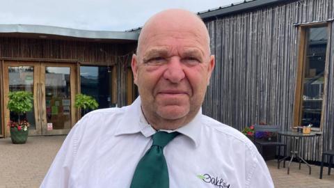 A bald man is wearing a white shirt and dark green tie. He is standing in front of a wood panelled building.