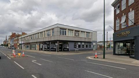 A road junction. A large building can be seen in the distance. A shop can be seen on the right of the road. The sky is overcast.