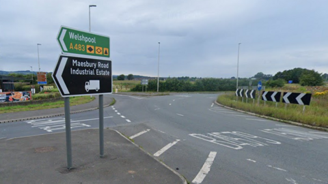 A roundabout with a green sign saying Welshpool and a black sign saying Maesbury Road industrial estate. White road markings can be seen on the roundabout.