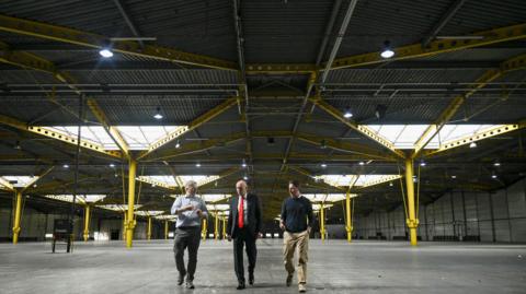 Three men walking through a large empty building space, which looks like the inside of a warehouse and has large yellow columns. 