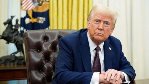 Donald Trump, wearing a blue suit with purple tie, sits at the Resolute Desk in the Oval Office