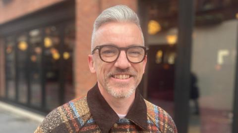 Johnny Rogan is wearing a coloured jacket with a brown collar. He has brown glasses on and short grey hair. Behind him is a blurry red brick building with black windows and doors.