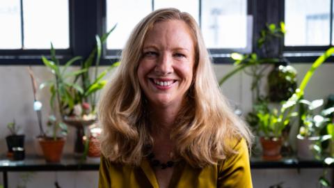 Karen Canfell, wearing a gold top, smiles at the camera, potted plants in the background behind her