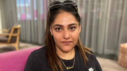 A woman looks directly into the camera - she has big brown eyes, a ponytail with long dark hair, pair of glasses on her head, a black t-shirt and gold necklace with the word 'Sahara', the name of her daughter who died.