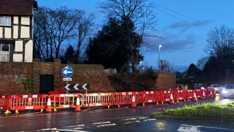 A photograph taken during low light with cars passing with their lights on. There is a red brick wall that has partially collapsed in the centre with orange plastic barriers covering up the area. 