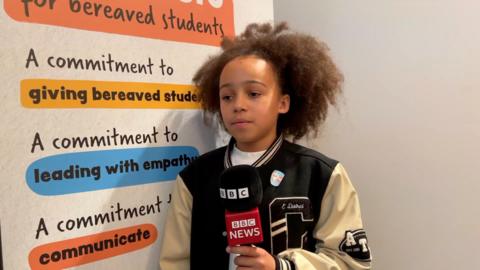 A child holds a microphone in front of a poster about bereavement. He has brown curly hair and wears a baseball jacket.