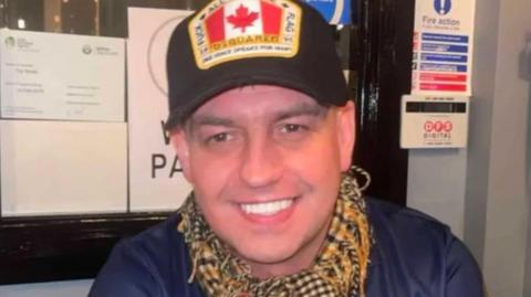 A man wearing a cap with a canadian flag on it, he is wearing a yellow check scarf. He is smiling