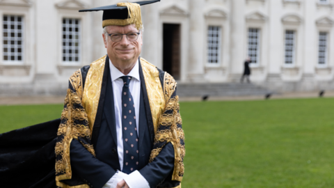 Chris Smith is wearing a black mortar board with gold trims and an ornate black and gold robe over a navy suit, white shirt and navy tie with emblems. He is standing up, with a lawn and grand building in the background.