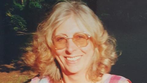 A faded image of a woman with curly blonde shoulder-length hair and tinted sunglasses smiling. Bright sun is shining in her face, and the woodland behind is partially in shadow. She is wearing a checked red and white dress.
