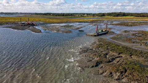  A shot of the restoration work being done at Lymington. Equipment can be seen set up on mud next to a body of water. 