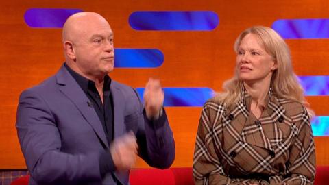 A bald man, Ross Kemp plays air drums while sitting next to Pamela Anderson, who has long blonde hair