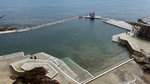 La Valette is an outdoor pool with a seawall enclosing some of the sea water. Steps lead down to the pool and there is some seaweed in the water. The coastline is just beyond.