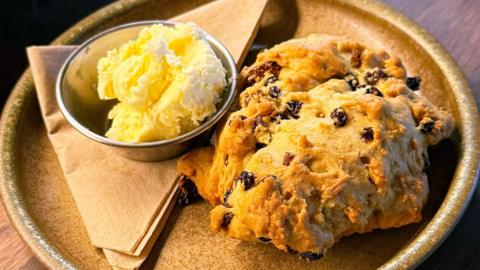A crumbly baked good with dried fruit on a plate next to small bowl of clotted cream.
© Nettle cafe