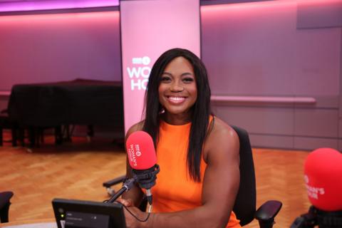 A woman with long, dark hair sits in front of a microphone and smiles at the camera. 